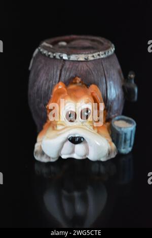 Statuette en céramique, une tirelire pour l'argent sous la forme d'un chien regardant hors d'une cabine en forme de tonneau avec de la bière sous la surface noire brillante. Banque D'Images