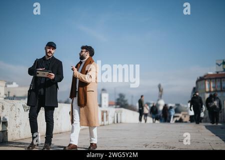 Une interaction professionnelle a lieu alors que deux hommes élégants s'engagent dans une conversation tout en se tenant sur un pont de la ville avec un ciel bleu clair au-dessus. Banque D'Images