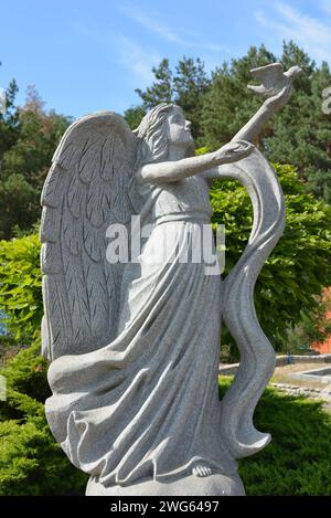 Statue en pierre d'un ange, une fille sur une pierre avec une colombe à la main sur un fond d'une forêt de pins et un ciel bleu. Banque D'Images