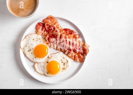 Petit déjeuner avec œufs au plat, bacon et café sur table blanche, vue de dessus, espace copie. Œufs de côté ensoleillés frits avec bacon sur l'assiette et cappuccino du matin Banque D'Images