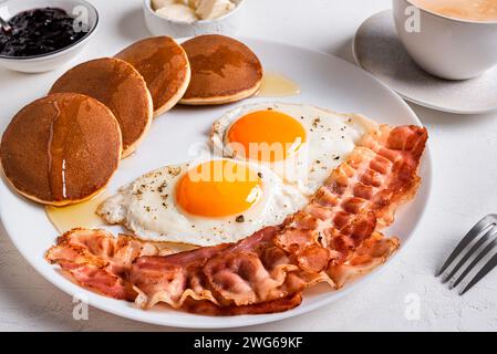 Petit déjeuner avec œufs au plat, bacon, crêpes et café sur la table blanche en gros plan. Œufs de côté ensoleillés frits avec bacon sur l'assiette et café cappuccino du matin Banque D'Images