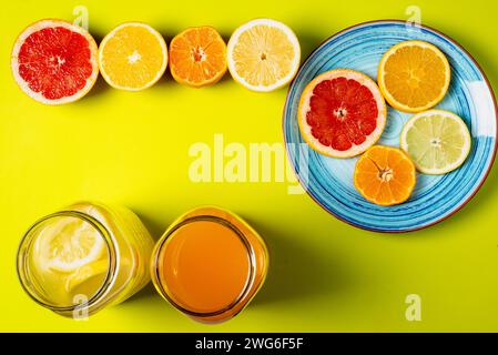Tranches de pamplemousse, orange, citron et mandarines sur une assiette avec demi-fruits et deux pots avec jus, fond verdâtre. Banque D'Images