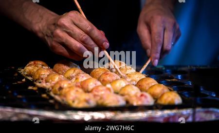 Un vendeur japonais prépare un Takoyaki sur la poêle chaude du Japon. Processus de cuisson des boules frites populaires avec poulpe dans la nourriture de rue locale. Faire un bal Banque D'Images