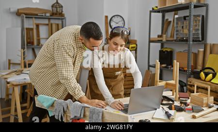 Un homme et une femme collaborent dans un atelier de menuiserie, examinant des conceptions sur un ordinateur portable entouré d'outils de travail du bois. Banque D'Images