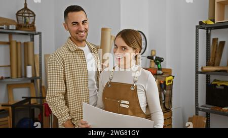 Un homme et une femme souriants debout dans un atelier de menuiserie lumineux tenant des plans et un rouleau de papier. Banque D'Images