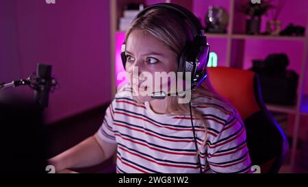 Femme caucasienne focalisée portant des écouteurs dans une salle de jeu colorée avec microphone et pc la nuit. Banque D'Images