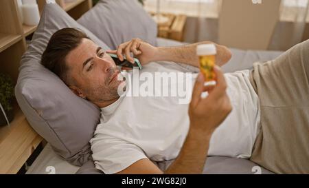 Jeune homme hispanique inquiet allongé malade dans son lit, serrant des pilules tout en ayant une conversation téléphonique sérieuse, engrossé dans sa chambre faiblement éclairée et confortable Banque D'Images