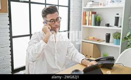 Jeune homme hispanique attrayant immergé dans des discussions d'affaires sérieuses sur smartphone tout en travaillant dur à l'intérieur au travail de bureau, concentré mais détendu prof Banque D'Images