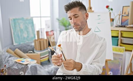 Jeune homme hispanique sérieux, un artiste émergent, profondément engagé dans le choix du pinceau parfait pour sa toile dans un studio d'art animé. Banque D'Images