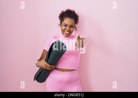Jeune femme hispanique avec les cheveux bouclés tenant tapis de yoga sur fond rose souriant amical offrant poignée de main comme salutation et accueil. réussi b Banque D'Images