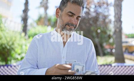 Un jeune homme hispanique confiant aux cheveux gris compte les billets d'un dollar, debout dans un parc ensoleillé, déclarant sa richesse avec un sourire joyeux. Banque D'Images