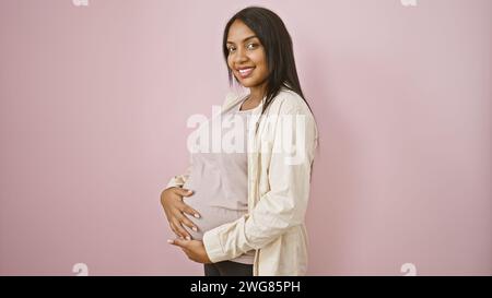 Jeune femme enceinte joyeuse avec un sourire radieux se réjouit de toucher joyeusement son ventre, debout avec désinvolture sur fond isolé rose Banque D'Images