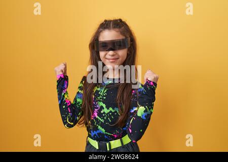 Adorable fille hispanique, immergé dans la victoire amusante des lunettes vr, crie dans l'excitation joyeuse! positivement exubérant, avec un geste gagnant contre isolé Banque D'Images