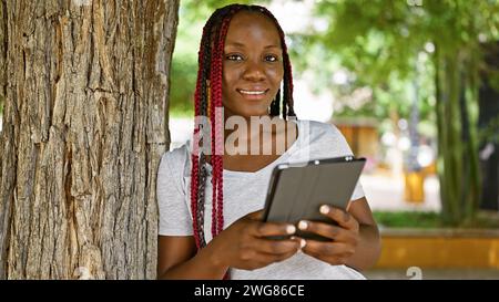 Femme afro-américaine confiante, décontracté élégant dans le parc de la ville, rayonne de positivité tout en utilisant le touchpad, son sourire prêtant un air de chee infectieux Banque D'Images