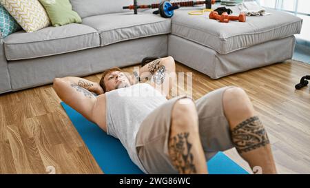 Un jeune homme, tatoué, barbu qui s'exerce à l'intérieur sur un tapis de yoga dans une pièce bien rangée avec un canapé gris et un équipement de fitness. Banque D'Images