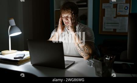 Jeune homme stressé avec la barbe ressentant des maux de tête la nuit au bureau. Banque D'Images