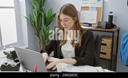 Une jeune femme caucasienne adulte travaille avec diligence dans un bureau intérieur, regardant concentré sur son ordinateur portable. Banque D'Images