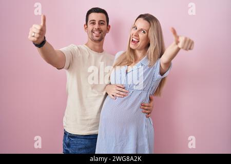 Jeune couple s'attendant à un bébé debout sur fond rose approuvant faire un geste positif avec la main, les pouces en souriant et heureux pour le succès. winne Banque D'Images