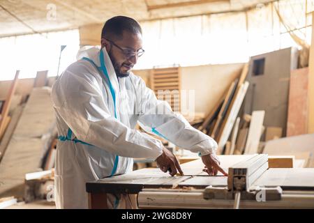 jeune homme avec des lunettes et des salopettes travaillant dans une usine de meubles, préparant des machines pour commencer à construire des meubles, le lieu de travail et le style de vie Banque D'Images