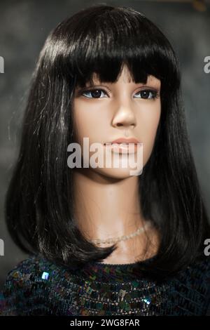 Mannequin femme en plastique avec de longs cheveux noirs portant un haut de paillettes posant sur un fond gris flou. Effet de lumière capillaire. Banque D'Images