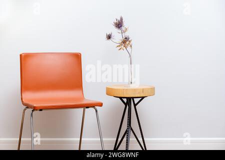 Un cadre minimaliste avec une chaise rouge élégante et une table ronde en bois avec un arrangement de fleurs séchées style Japandi dans un vase clair, contre un sof Banque D'Images