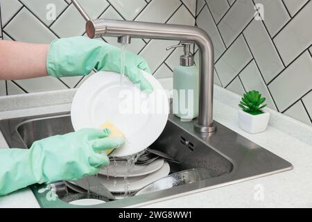 Gros plan des mains dans des gants verts lavant une assiette blanche avec une éponge sous l'eau courante du robinet, pile de vaisselle sale ou des assiettes dans le robinet de cuisine Banque D'Images