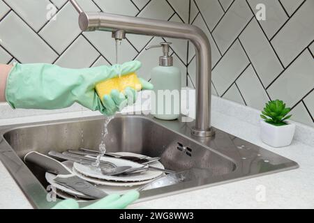 Main dans le gant vert presse l'éponge sous le robinet. Assiettes et couverts attendent le nettoyage dans l'évier hygiène domestique, corvées Banque D'Images