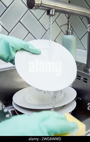 Gros plan des mains dans des gants verts lavant une assiette blanche avec une éponge sous l'eau courante du robinet, pile de vaisselle sale ou des assiettes dans le robinet de cuisine Banque D'Images