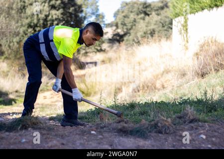 Vue latérale d'un travailleur municipal en uniforme utilisant une houe de fer pour enlever l'herbe séchée à l'extérieur Banque D'Images