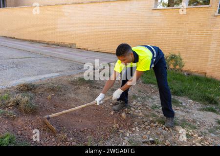 Vue latérale d'un travailleur municipal en uniforme utilisant une houe de fer pour enlever l'herbe séchée à l'extérieur Banque D'Images