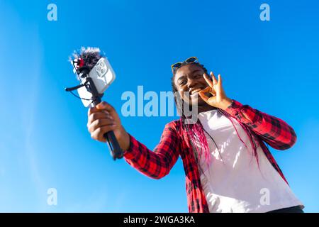 Photo vue à angle bas d'une femme africaine souriante gestionnant le succès lors d'un streaming en ligne sous le soleil dans une journée d'été Banque D'Images