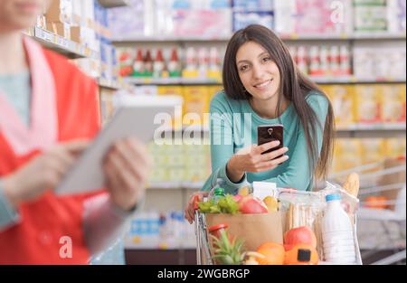 Jeune femme utilisant un smartphone au supermarché et souriant à la caméra Banque D'Images