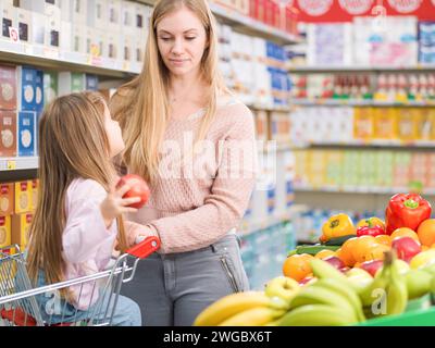 Heureuse fille mignonne assise sur un panier et achetant des légumes frais et des fruits avec sa mère au supermarché Banque D'Images