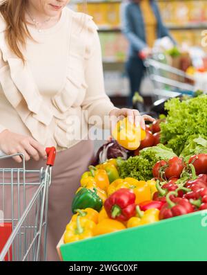 Femme poussant un chariot et achetant des légumes frais au supermarché, elle cueille un poivron Banque D'Images