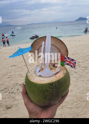 Phuket, Thaïlande - 24 décembre 2023 : cocktail rafraîchissant de noix de coco sur l'île de Khai Nai dans les mains de l'homme sur la plage Banque D'Images