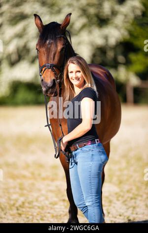 Une jeune femme se tient debout avec son cheval sur une arène d'équitation ensoleillée. En arrière-plan, arbres en fleurs et nature verte, modèle isolé devant le flou, bro Banque D'Images