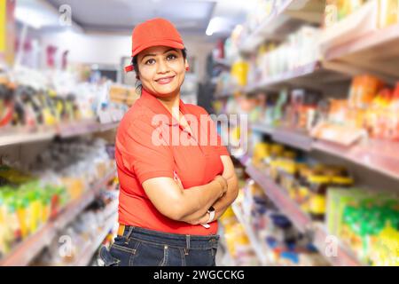 Heureux jeune indien personnel de magasin d'épicerie portant T-shirt rouge et casquette debout avec des bras croisés dans le supermarché. Concept d'autonomisation des femmes Banque D'Images