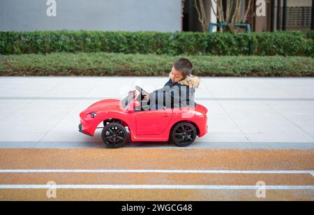 Un jeune enfant multiracial navigue joyeusement sur la piste de course du quartier dans une voiture de sport rouge, enfilant un grand sourire alors qu'ils prennent le contrôle du Banque D'Images