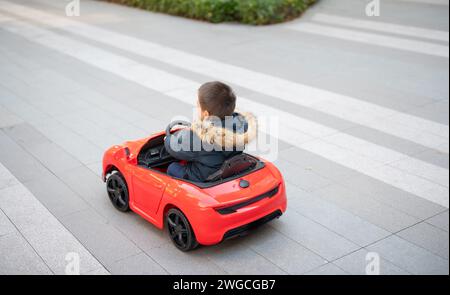 Un jeune enfant multiracial navigue joyeusement la route du quartier dans une voiture de sport rouge jouet, enfilant un grand sourire alors qu'ils prennent le contrôle de leur jouet ca Banque D'Images