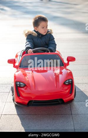 Un jeune enfant multiracial navigue joyeusement la route du quartier dans une voiture de sport rouge jouet, enfilant un grand sourire alors qu'ils prennent le contrôle de leur jouet ca Banque D'Images