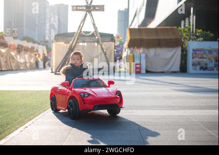 Un jeune enfant multiracial navigue joyeusement la route du quartier dans une voiture de sport rouge jouet, enfilant un grand sourire alors qu'ils prennent le contrôle de leur jouet ca Banque D'Images