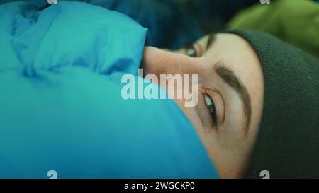 La personne se réveille dans le sac de couchage de tente de camping de près. Portrait de jeune fille touristique ouvrant les yeux dans le matin froid. Vacances femme voyageuse, trekking, randonnée, tourisme et activité de plein air. Vue latérale Banque D'Images