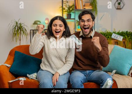 Heureux jeune couple indien avec des poings de mains levantes criant, célébrant le succès gagnant de la loterie jackpot réalisation d'objectif, bonne nouvelle victoire. Argent cash dollar pluie tombant sur la famille sur le canapé à la maison. Banque D'Images