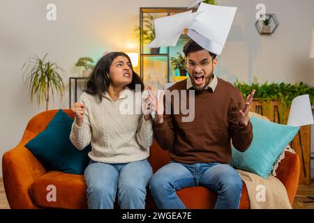 Stressé jeune couple diversifié regardant les factures de dette bancaire impayées, faire de la paperasse, planifier le budget, calculer les paiements hypothécaires des finances. La famille bouleversée ensemble jette des factures de papier maison assis sur le canapé. Banque D'Images