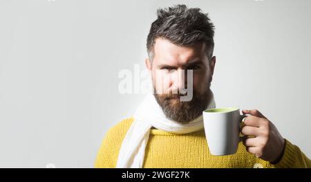 Homme sérieux en pull tricoté enveloppé dans une écharpe avec une tasse de café ou de thé. Automne ou hiver. Bel homme barbu dans des vêtements chauds buvant Banque D'Images