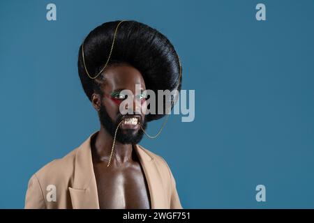Portrait d'homme gay afro-américain avec maquillage et perruque vintage noire. Personne LGBT tenant un accessoire en or avec des dents et portant une veste sur fond bleu. Banque D'Images