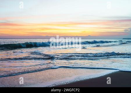 Le soleil se couche sur la plage tranquille, projetant des teintes chaudes à travers le ciel et reflétant sa lueur rose et dorée sur les vagues qui se détendent doucement Banque D'Images