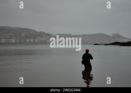 Baiona, Pontevedra, Espagne ; 12 juillet 2021 : sur la plage de Ladeira, un pêcheur tente de pêcher par une journée brumeuse avec Baiona en arrière-plan et au genou profond dans le se Banque D'Images