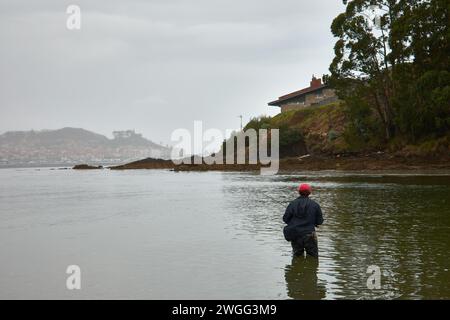 Baiona, Pontevedra, Espagne ; 12 juillet 2021 : sur la plage de Ladeira, un pêcheur tente de pêcher par une journée brumeuse avec Baiona en arrière-plan et au genou profond dans le se Banque D'Images