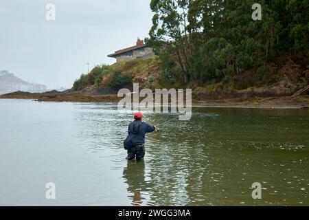 Baiona, Pontevedra, Espagne ; 12 juillet 2021 : sur la plage de Ladeira, un pêcheur tente de pêcher par une journée brumeuse avec Baiona en arrière-plan et au genou profond dans le se Banque D'Images